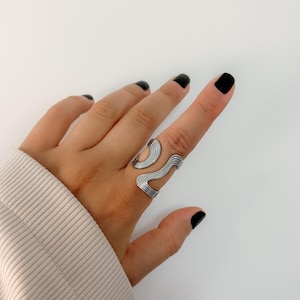 May include: Silver-toned ring with a wavy, abstract design. The ring features a series of curved lines and a smooth, polished finish. The ring is worn on a finger, showcasing its unique, modern style. The ring is a piece of jewelry.