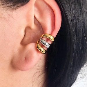 May include: A gold and silver ear cuff. The ear cuff has a layered design with two gold bands and one silver band. The jewelry is worn on the ear. The ear cuff is a modern and stylish accessory.