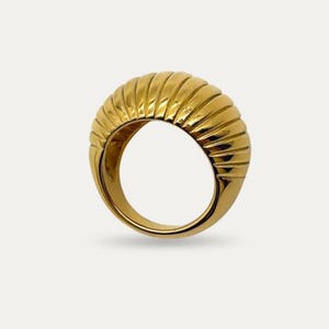 May include: A gold ring featuring a ribbed, rounded top. The ring has a smooth band and a textured, ridged design on the top, creating a bold, sculptural appearance. The ring is set against a white background.
