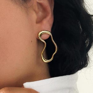 May include: A gold-colored, abstract-shaped earring. The earring has a fluid, organic design with smooth, rounded edges. The earring is worn on the ear, and the background is blurred.