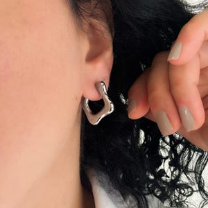 May include: A close-up of a silver earring with an irregular, organic shape. The earring is worn on the ear, and the design features a flowing, abstract form. The jewelry is shiny and reflects light.