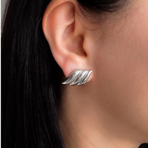 May include: A silver-toned ear cuff earring with a layered, wave-like design. The earring is worn on the ear, showcasing its textured, metallic surface. The jewelry piece has a modern, minimalist aesthetic.