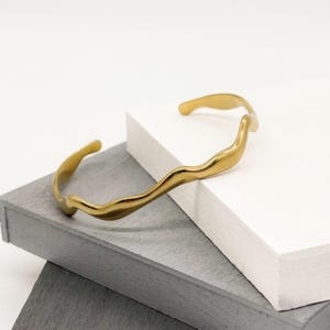 May include: A gold-colored cuff bracelet with a flowing, organic design. The open-ended bracelet has a wavy, textured surface, creating a fluid, sculptural aesthetic. The bracelet is displayed on a white and gray surface.