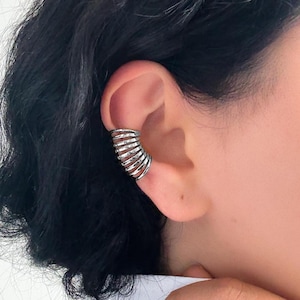 May include: Silver ear cuff featuring a layered, spiral design. The cuff wraps around the ear cartilage, creating a modern and edgy look. The jewelry is shown on a person's ear, highlighting its intricate details and metallic shine.