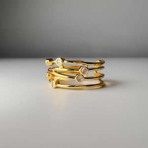 May include: A gold-toned multi-band ring featuring a layered, overlapping design. The ring is constructed from several thin, polished gold-colored metal wires that wrap around the finger, accented with four small, clear, round-cut stones.