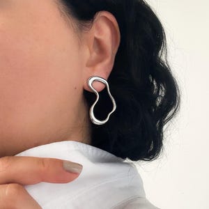 May include: A single silver earring with an abstract, flowing design is shown on an ear. The earring has a smooth, polished surface and an organic shape. The background includes a white shirt and dark hair. The earring's design is modern.