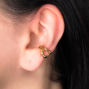 May include: A gold-colored ear cuff is worn on the ear. The ear cuff has a double-band design that curves around the ear. The ear cuff is made of a shiny, metallic material. The background is a close-up of the ear and dark hair.