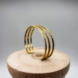 May include: A gold-colored cuff bracelet with three parallel bands. The bands have a textured surface and are open at one end. The bracelet is displayed on a wooden stand against a neutral background.