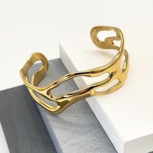 May include: A gold-colored cuff bracelet with an open, flowing design. The bracelet has an organic shape with multiple cut-out sections. The metal has a polished, reflective surface, and the bracelet is displayed on a white and gray surface.