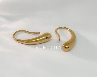 May include: Two gold-coloured teardrop earrings. The earrings have a smooth, polished finish and a curved design. The word "ELETTRA" is printed in white on the bottom of the image. The earrings are displayed on a white background.