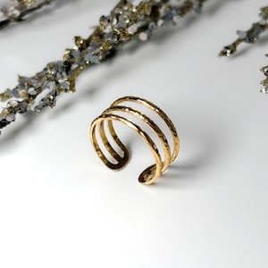 May include: A gold-toned ring featuring three parallel bands connected at the back, creating an open-style design. The surface of the metal bands has a textured, hammered finish that catches the light, providing a reflective appearance.