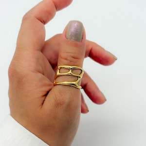 May include: A gold-colored ring with an abstract, open design. The ring features a layered structure with irregular, organic shapes. The ring is worn on a finger, showcasing its bright, reflective surface. The jewelry piece is a statement accessory.