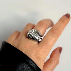 May include: A silver ring with a ribbed, rounded design. The ring is worn on a finger, and the design features parallel, curved lines. The ring's surface reflects light, creating highlights and shadows. The ring is a piece of jewellery.