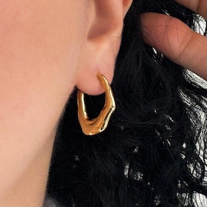 May include: A close-up of a gold-colored hoop earring with an irregular, organic shape. The earring is worn on a person's ear, with dark hair visible in the background. The earring has a smooth, reflective surface.