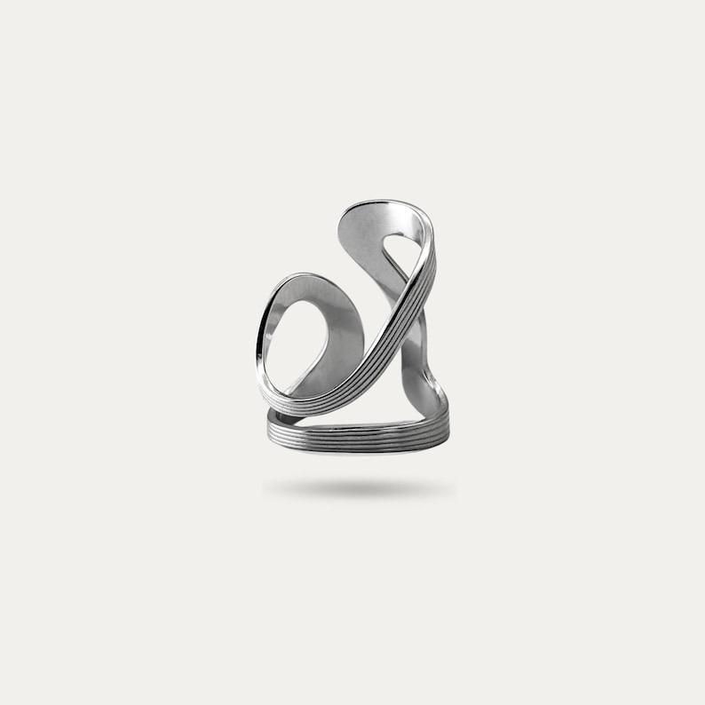 May include: A silver-colored ring with an abstract design. The ring features a looping, flowing shape with multiple bands and curves. The design is modern and sculptural, with a polished finish. The ring appears to be made of metal.