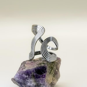 May include: A silver-toned, abstract ring with a layered, wave-like design. The ring is displayed on a rough, purple and gray stone, set against a neutral background. The ring's design features multiple parallel lines, creating a textured appearance.