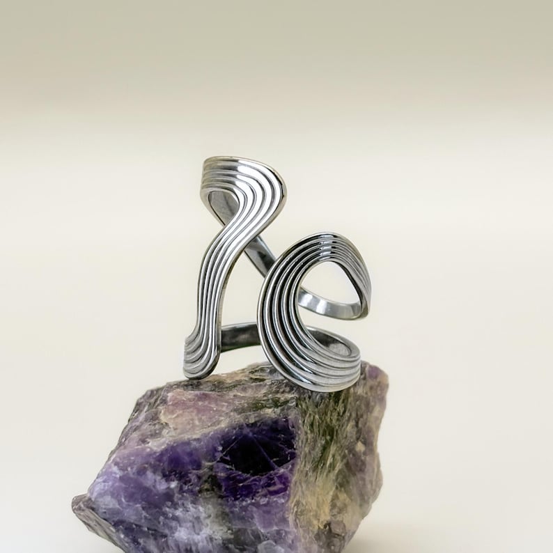 May include: A silver-toned, abstract ring with a layered, wave-like design. The ring is displayed on a rough, purple and gray stone, set against a neutral background. The ring's design features multiple parallel lines, creating a textured appearance.