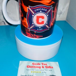 Puede incluir: Taza de cerámica blanca con un diseño de un logotipo de Chicago Fire rojo y azul con llamas en el fondo. La taza está sobre un posavasos azul y blanco. La imagen también incluye una tarjeta de presentación para Geek-Tee Clothing & Gifts.
