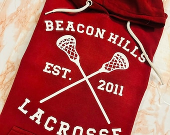 May include: Red hooded sweatshirt with white text and lacrosse stick graphic. The text reads "Beacon Hills Est. 2011 Lacrosse".