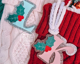 May include: Two Christmas stocking ornaments. One is light blue, the other is pink, both with a holly leaf design and silver glitter trim. The ornaments are hanging on white and red knitted stockings.