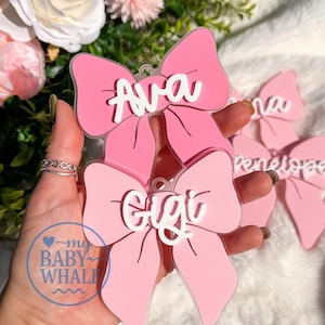 May include: Two pink acrylic bow-shaped ornaments featuring white raised lettering. The top bow displays the name Ava, and the bottom bow displays the name Gigi. Each bow has a small circular hole at the top for hanging, with a clear acrylic base.