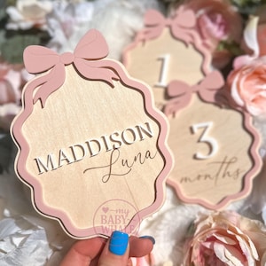 May include: Wooden baby milestone markers with a pink bow design. One marker has the name "Maddison Luna" engraved. Another marker shows the number "1". The third marker shows the number "3" and the word "months". The markers are surrounded by flowers.