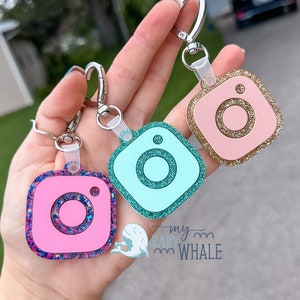 May include: Three Instagram-themed keychains with glitter accents. The keychains are pink, blue, and gold. The keychains are attached to silver key rings.