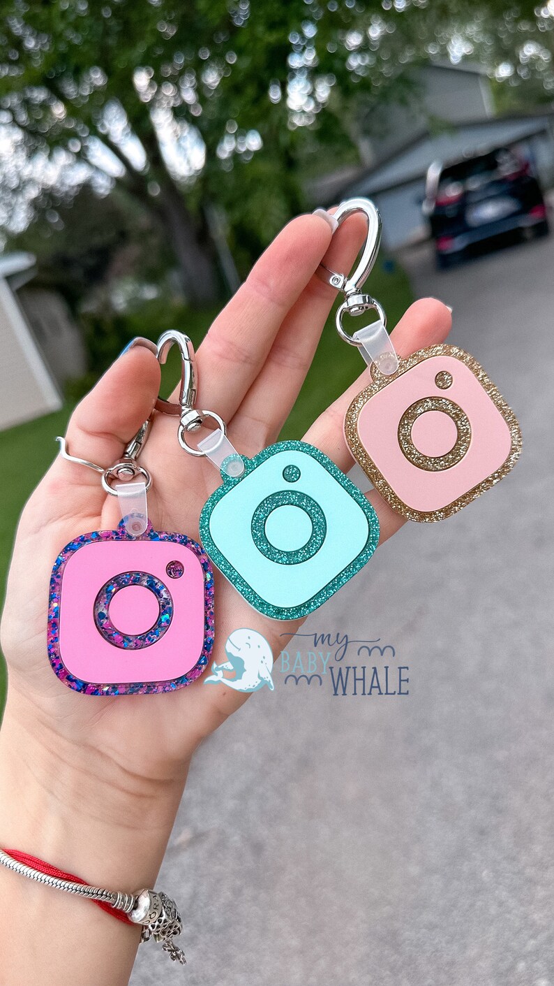 May include: Three Instagram-themed keychains with glitter accents. The keychains are pink, blue, and gold. The keychains are attached to silver key rings.