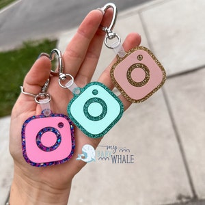 May include: Three Instagram-themed keychains with glitter accents. The keychains are pink, teal, and gold. Each keychain features a silver ring and a clear plastic loop.