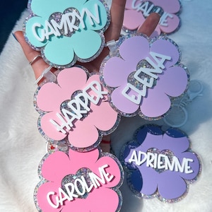 K&ouml;nnte beinhalten: Eine Sammlung von bl&uuml;tenf&ouml;rmigen Schl&uuml;sselanh&auml;ngern in Pastellfarben wie Rosa, Blau und Lila. Jeder Schl&uuml;sselanh&auml;nger tr&auml;gt einen Namen in wei&szlig;er Schrift: Camryn, Carrie, Harper, Elena, Caroline und Adrienne. Die R&auml;nder der Bl&uuml;ten sind mit Glitzer versehen.