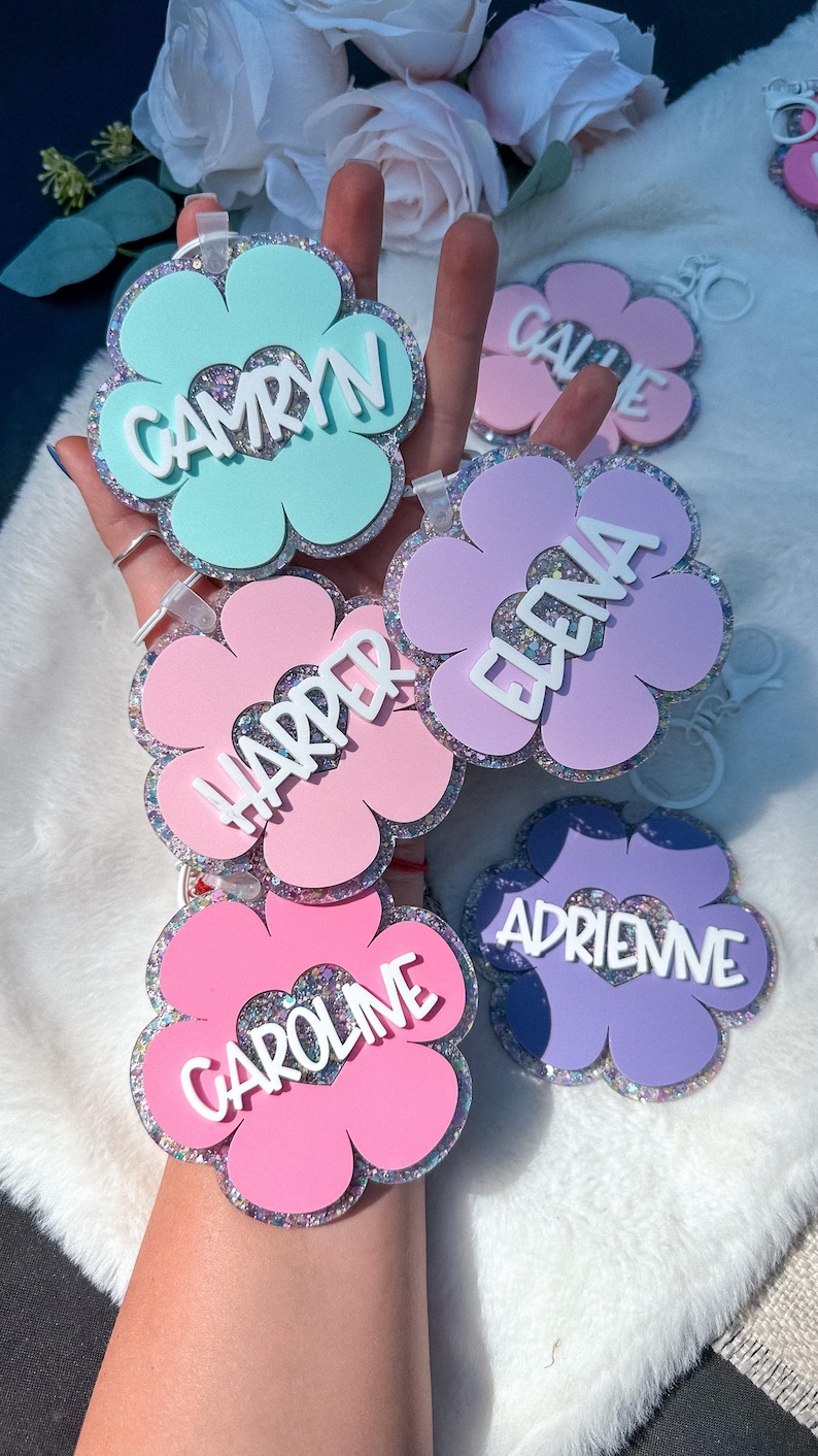 K&ouml;nnte beinhalten: Eine Sammlung von bl&uuml;tenf&ouml;rmigen Schl&uuml;sselanh&auml;ngern in Pastellfarben wie Rosa, Blau und Lila. Jeder Schl&uuml;sselanh&auml;nger tr&auml;gt einen Namen in wei&szlig;er Schrift: Camryn, Carrie, Harper, Elena, Caroline und Adrienne. Die R&auml;nder der Bl&uuml;ten sind mit Glitzer versehen.