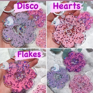 K&ouml;nnte beinhalten: Vier bl&uuml;tenf&ouml;rmige Schl&uuml;sselanh&auml;nger mit Glitzer und Herz- oder Flockenakzenten. Die Schl&uuml;sselanh&auml;nger sind in Rosa- und Lilat&ouml;nen gehalten. Der Text "Disco", "Hearts" und "Flakes" ist auf dem Bild zu sehen. Jeder Schl&uuml;sselanh&auml;nger hat einen silberfarbenen Ring.