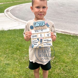 May include: A young child holding a wooden sign that says "CARTER" and "TODAY IS MY LAST DAY of Pre-K." The sign includes the date 5/27/25, age 5, and a time of 45 minutes. The child is wearing a tan shirt, black shorts, and sneakers.