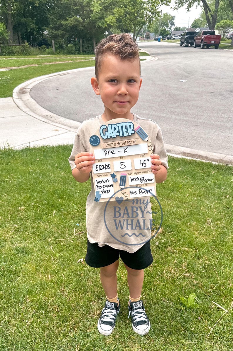 May include: A young child holding a wooden sign that says "CARTER" and "TODAY IS MY LAST DAY of Pre-K." The sign includes the date 5/27/25, age 5, and a time of 45 minutes. The child is wearing a tan shirt, black shorts, and sneakers.