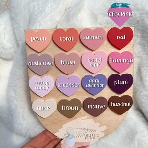 May include: A wooden color chart featuring heart-shaped color swatches. The chart includes a variety of colors such as peach, coral, salmon, red, dusty rose, blush, pastel pink, flamingo, cotton candy, lavender, dark lavender, plum, nude, brown, mauve, and hazelnut.