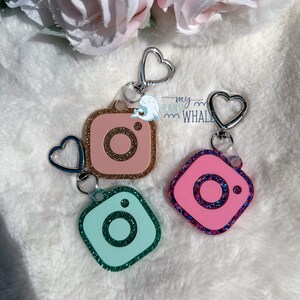 May include: Three sparkly Instagram camera keychains with silver heart-shaped rings. The keychains are pink, light blue, and dark pink with glitter.
