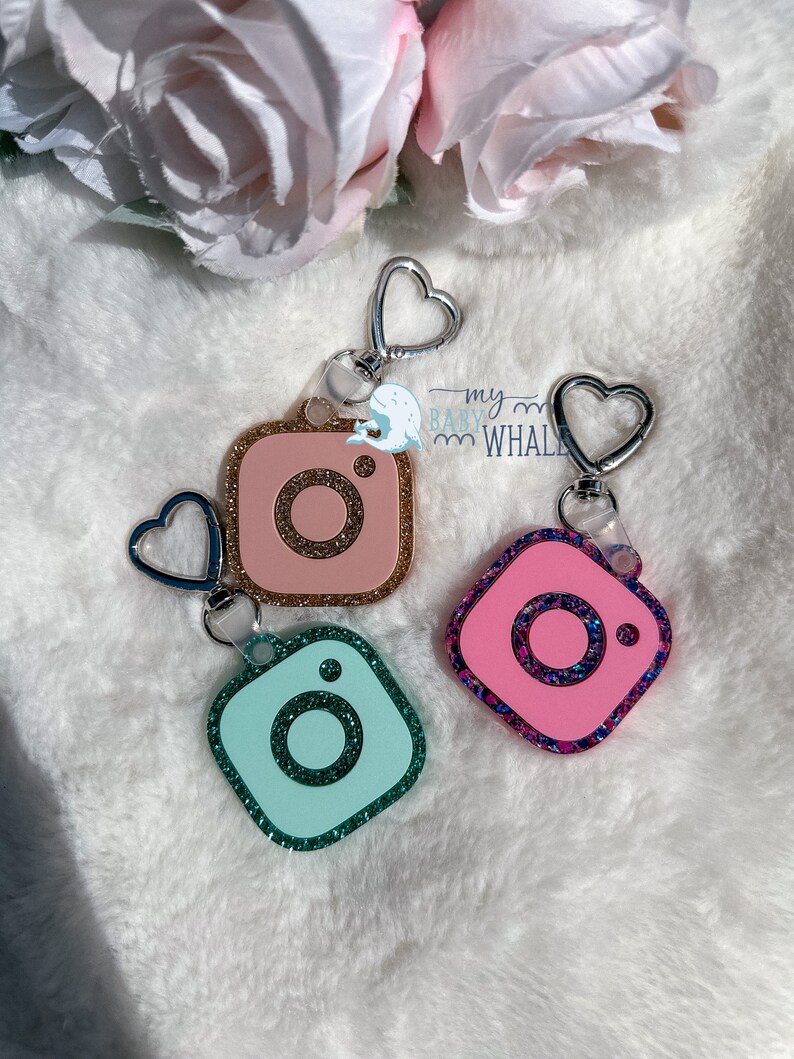 May include: Three sparkly Instagram camera keychains with silver heart-shaped rings. The keychains are pink, light blue, and dark pink with glitter.