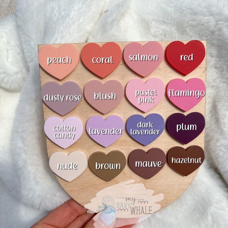 May include: A wooden board with 16 heart-shaped silicone pieces in various shades of pink, purple, and brown. Each heart has a different color name written on it, such as "peach", "coral", "salmon", "red", "dusty rose", "blush", "pastel pink", "flamingo", "cotton candy", "lavender", "dark lavender", "plum", "nude", "brown", "mauve", and "hazelnut".