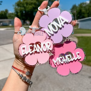 K&ouml;nnte beinhalten: Blumenf&ouml;rmige Schl&uuml;sselanh&auml;nger in Rosa und Lavendel mit Glitzerakzenten. Jeder Schl&uuml;sselanh&auml;nger hat einen Namen in wei&szlig;en Buchstaben: NOVA, ELEANOR und MICHELLE. Die Schl&uuml;sselanh&auml;nger werden vor einem hellen, sonnigen Hintergrund gehalten.