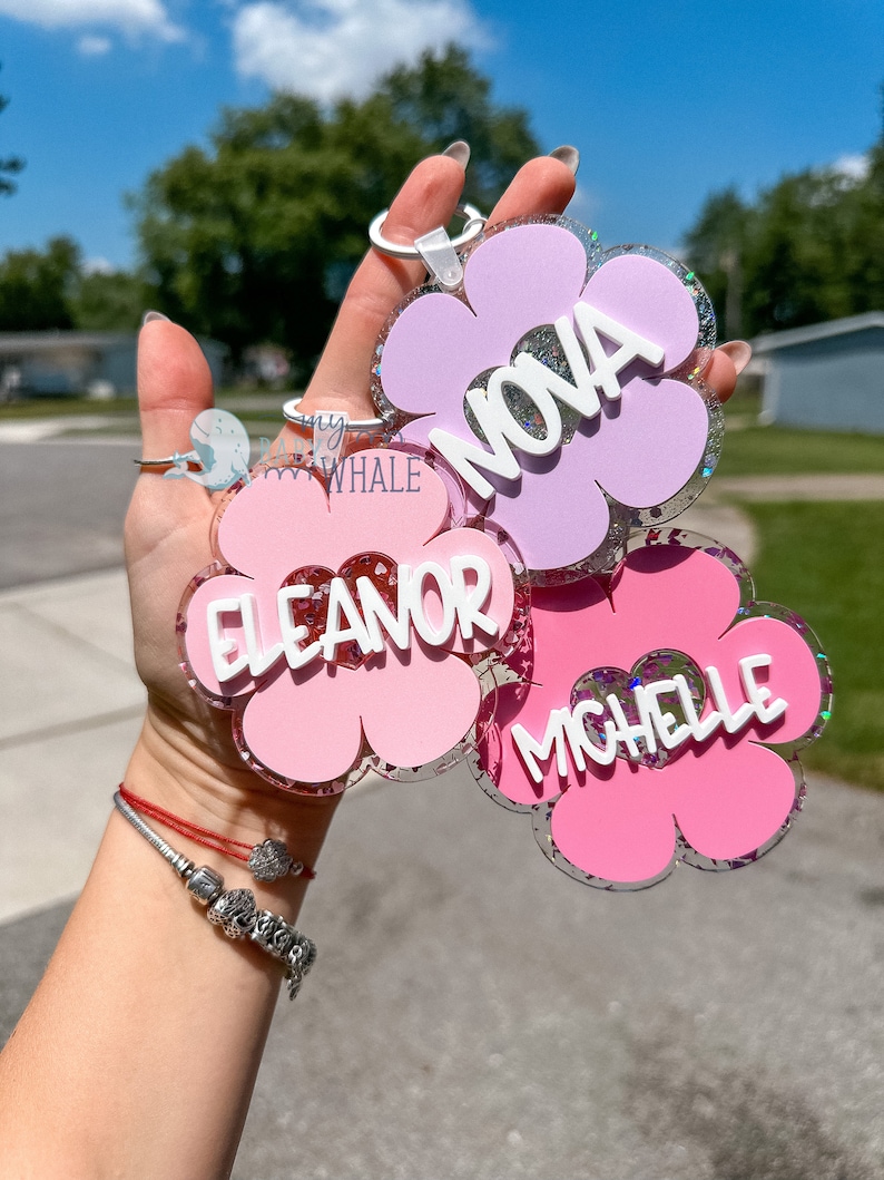 K&ouml;nnte beinhalten: Blumenf&ouml;rmige Schl&uuml;sselanh&auml;nger in Rosa und Lavendel mit Glitzerakzenten. Jeder Schl&uuml;sselanh&auml;nger hat einen Namen in wei&szlig;en Buchstaben: NOVA, ELEANOR und MICHELLE. Die Schl&uuml;sselanh&auml;nger werden vor einem hellen, sonnigen Hintergrund gehalten.