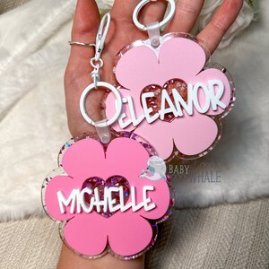 K&ouml;nnte beinhalten: Zwei rosa bl&uuml;tenf&ouml;rmige Schl&uuml;sselanh&auml;nger mit wei&szlig;en Akzenten und den Namen "Eleanor" und "Michelle" in wei&szlig;er Schrift. Die Schl&uuml;sselanh&auml;nger haben eine herzf&ouml;rmige Mitte, die mit Glitzer gef&uuml;llt ist, und sind an wei&szlig;en Schl&uuml;sselringen befestigt.