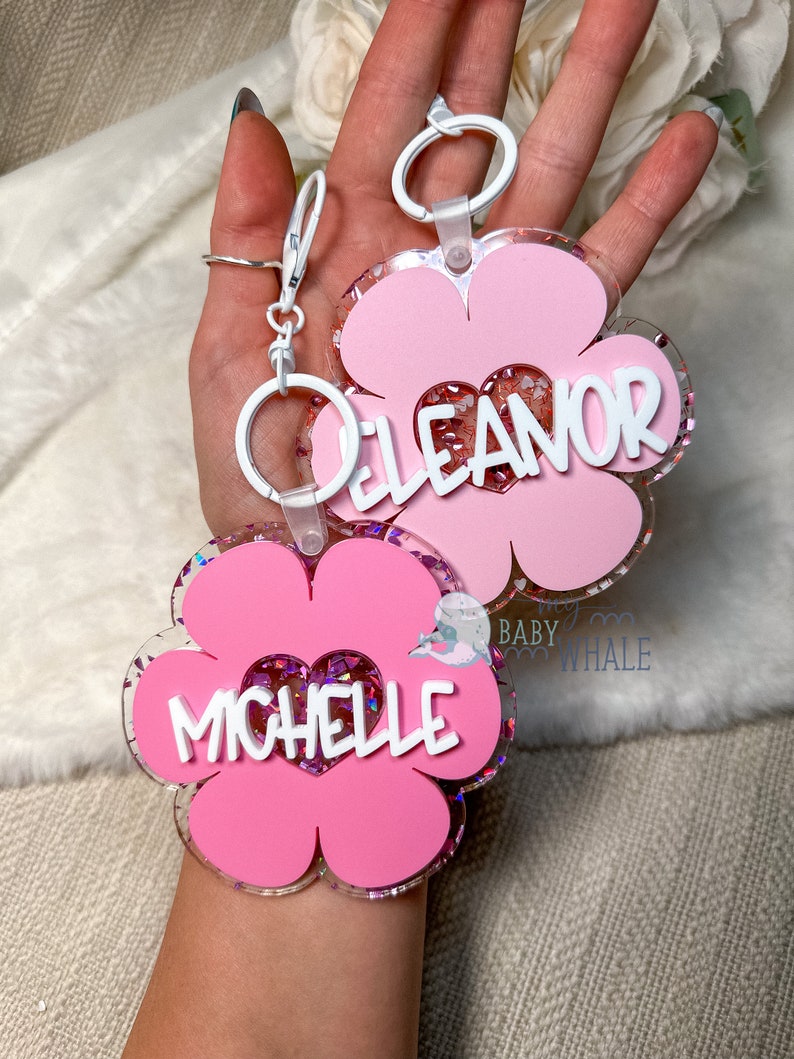 K&ouml;nnte beinhalten: Zwei rosa bl&uuml;tenf&ouml;rmige Schl&uuml;sselanh&auml;nger mit wei&szlig;en Akzenten und den Namen "Eleanor" und "Michelle" in wei&szlig;er Schrift. Die Schl&uuml;sselanh&auml;nger haben eine herzf&ouml;rmige Mitte, die mit Glitzer gef&uuml;llt ist, und sind an wei&szlig;en Schl&uuml;sselringen befestigt.