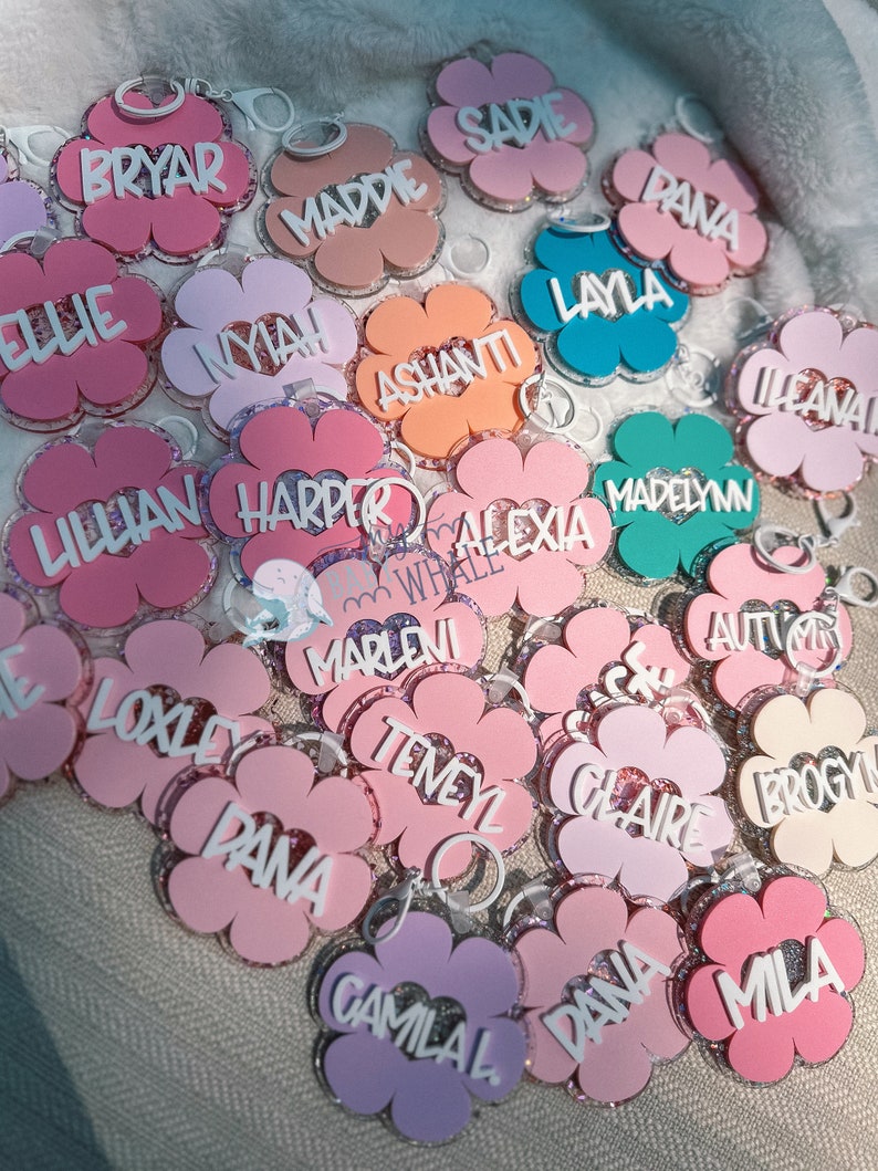 K&ouml;nnte beinhalten: Eine Sammlung von bl&uuml;tenf&ouml;rmigen Schl&uuml;sselanh&auml;ngern in verschiedenen Rosa-, Lila- und Oranget&ouml;nen. Jeder Schl&uuml;sselanh&auml;nger tr&auml;gt einen Namen in wei&szlig;er Schrift. Die Schl&uuml;sselanh&auml;nger haben einen glitzernden Rand und einen silbernen Ring.