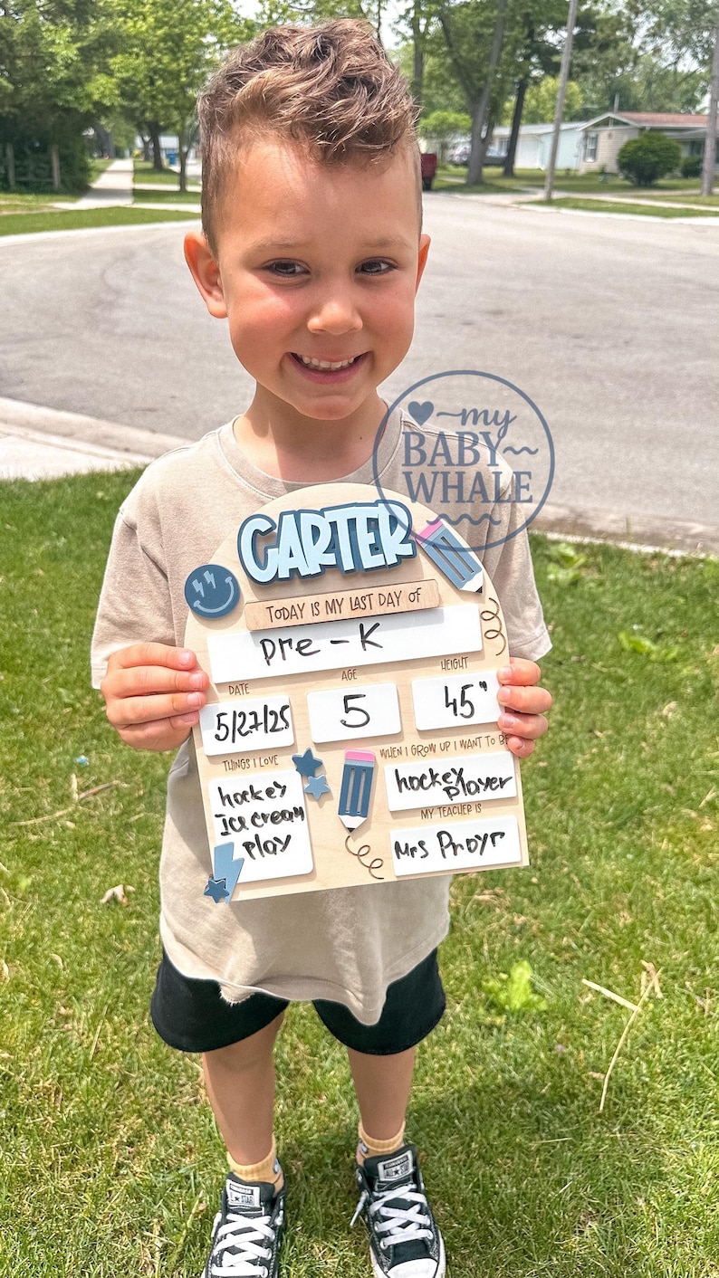 May include: A young child holds a wooden sign with the name "CARTER" and details about their last day of Pre-K. The sign includes the date, age 5, and height 45 inches. The child is wearing a beige shirt and black shorts.
