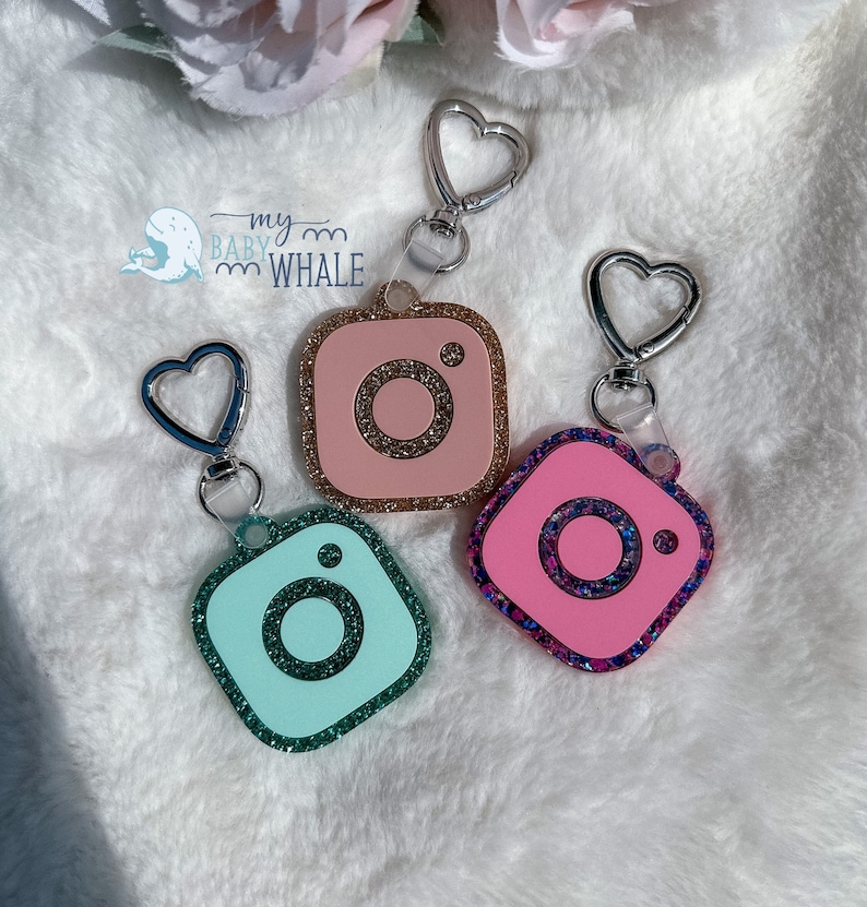 May include: Three Instagram logo keychains with glitter accents. The keychains are pink, blue, and green. Each keychain has a silver heart-shaped clasp.