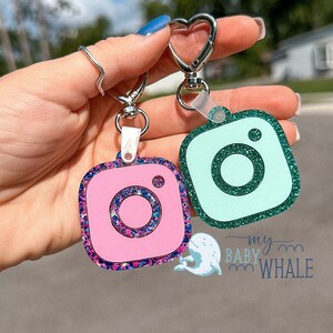 May include: Two Instagram camera keychains, one pink with blue and purple glitter and one teal with green glitter. The keychains are attached to a silver heart-shaped keyring.