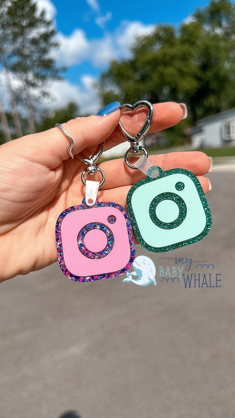 May include: Two Instagram camera keychains, one pink with blue and purple glitter and one teal with green glitter. The keychains are attached to a silver heart-shaped keyring.