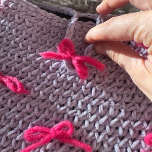 May include: A close-up of a lavender crocheted fabric with pink fuzzy bows attached. The fabric has a woven pattern.