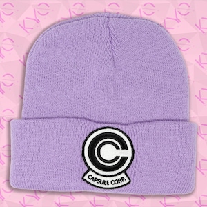 May include: A lavender beanie with a folded brim. The beanie features a black and white Capsule Corp. logo patch on the front. The hat is made of a knitted material.