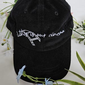 Puede incluir: Gorra de b&eacute;isbol de pana negra con bordado blanco que dice "This is our cinema" y un gr&aacute;fico de carrete de pel&iacute;cula.